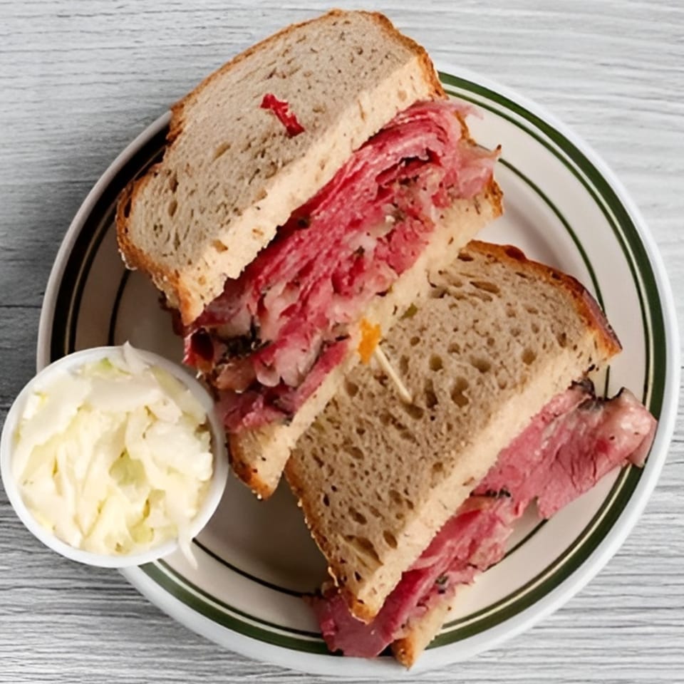 A pastrami sandwich on rye bread with a side of coleslaw on a white plate.