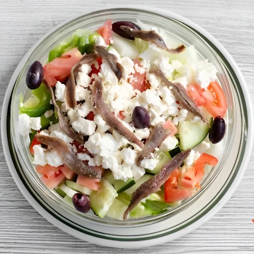 A fresh salad with lettuce, tomatoes, cucumbers, feta cheese, olives, and anchovies in a glass bowl.