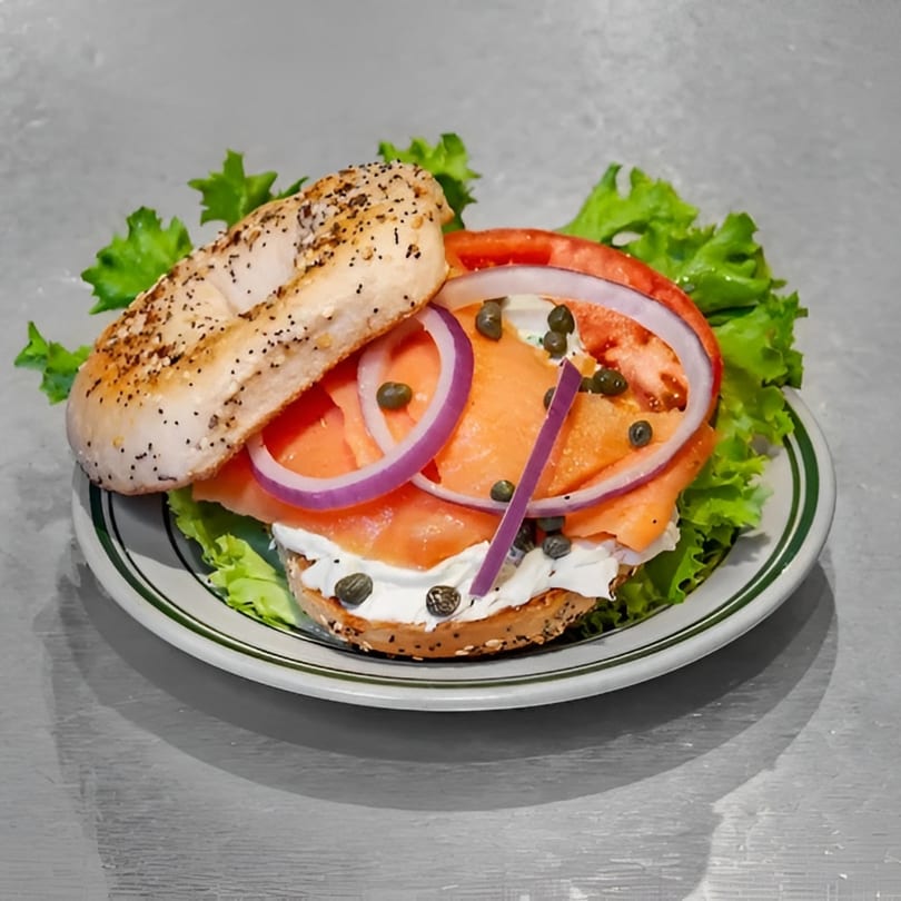 A poppy seed bagel with lox, cream cheese, red onion, capers, and lettuce on a plate.