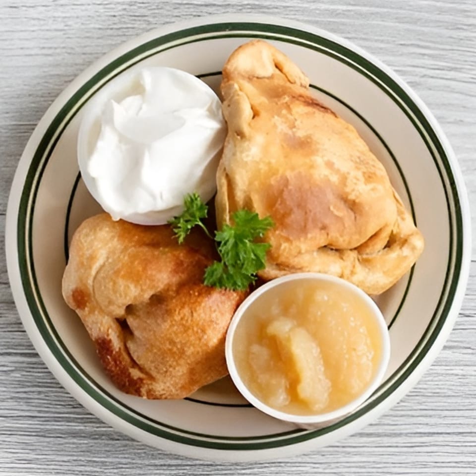 Two golden brown knishes with a side of sour cream and applesauce, garnished with parsley.