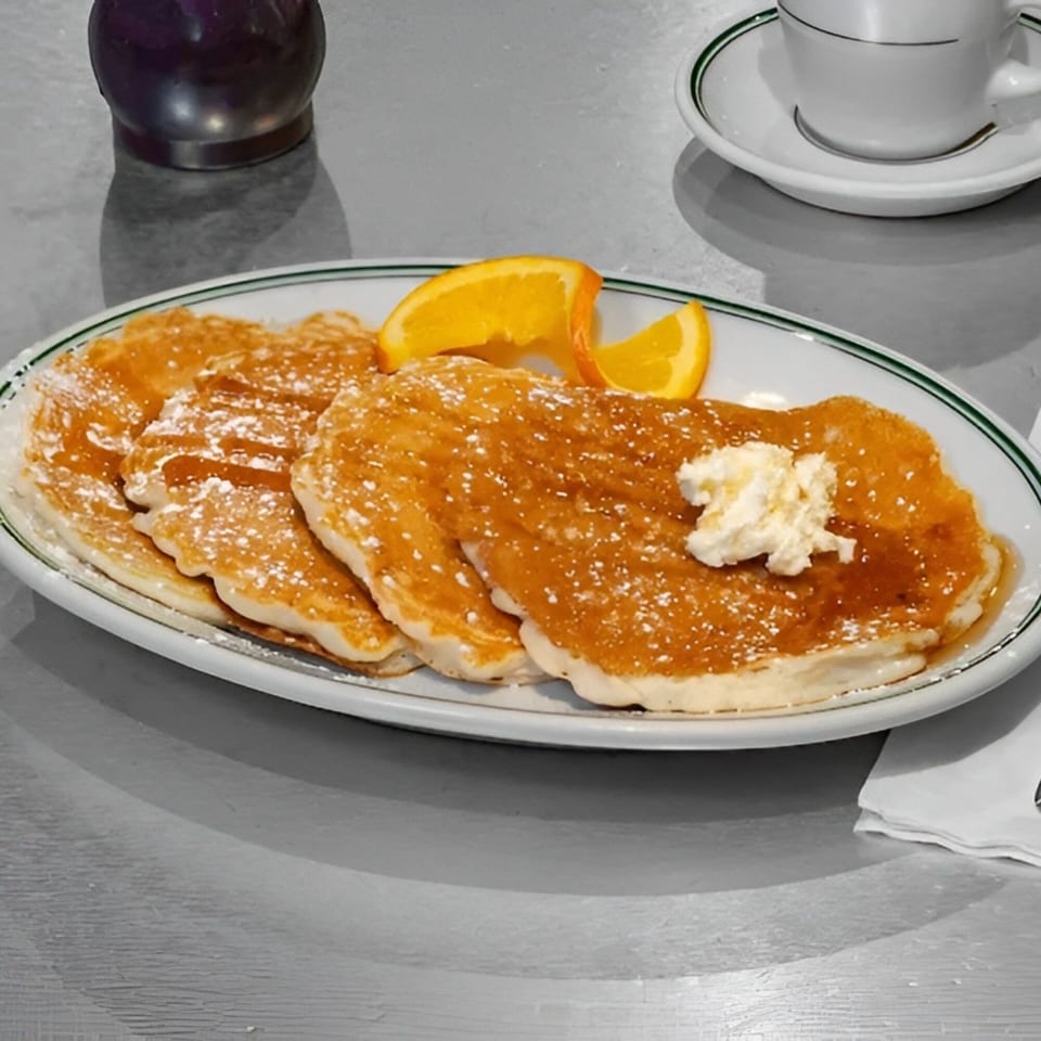 A plate of golden pancakes with syrup, powdered sugar, butter, and orange slices, next to a coffee cup.