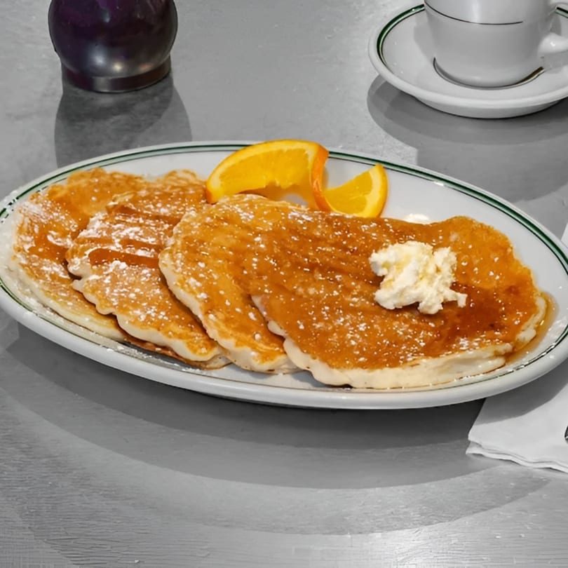 A plate of golden pancakes with syrup, powdered sugar, butter, and orange slices, next to a coffee cup.